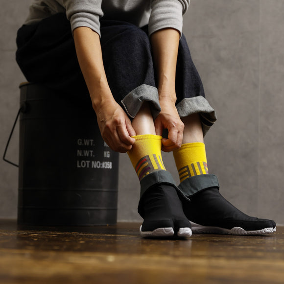 Person wearing yellow Tabi socks “Sankuzushi” with black Tabi shoes “RAIN,” adjusting the socks while sitting on a metal container.